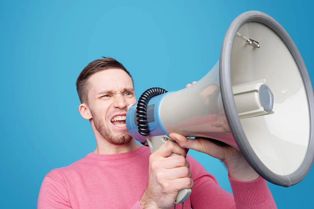 A man in a pink jumper shouting into a large megaphone against a bright blue background