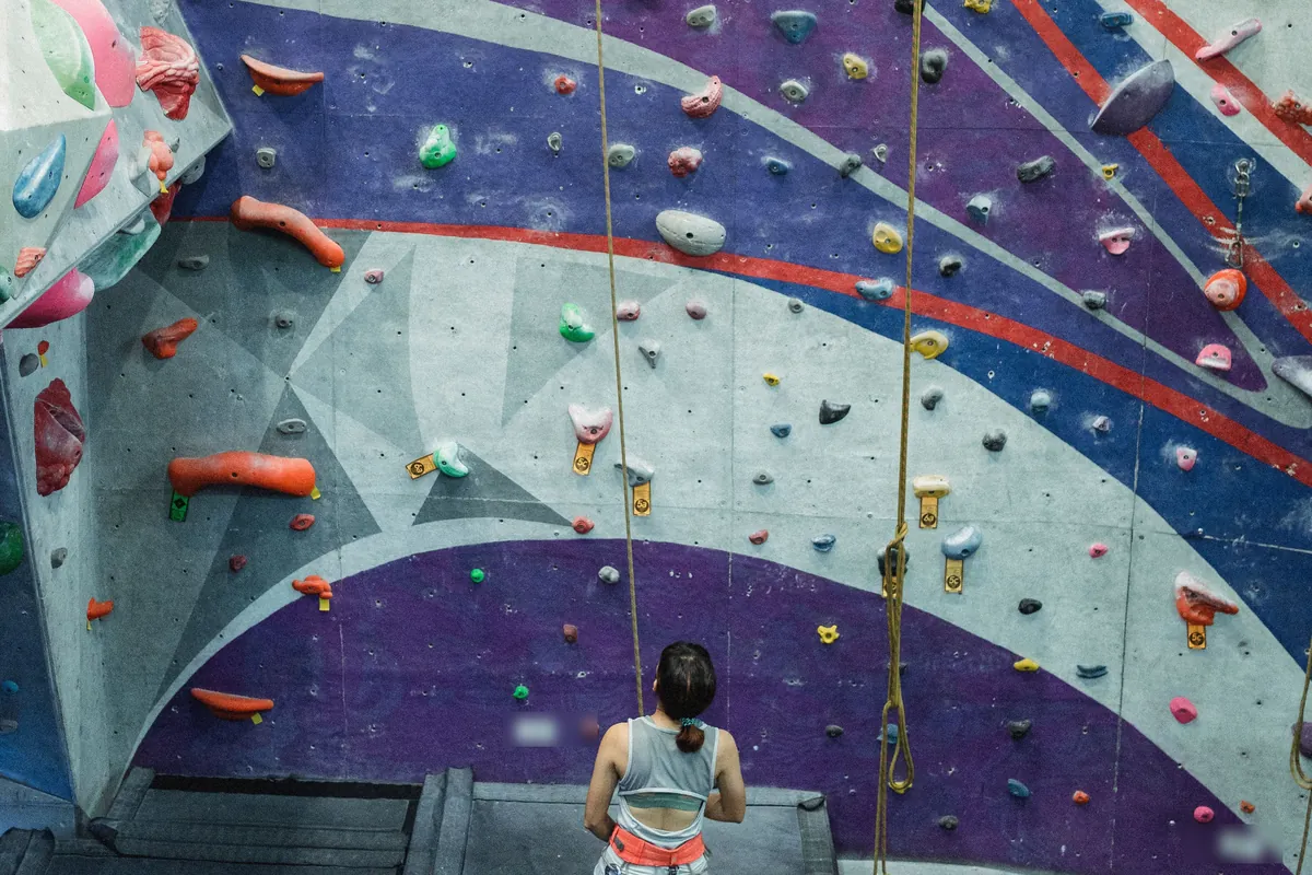 A climber’s partner holds the rope and looks up at a climbing wall painted in purple, blue and red bands