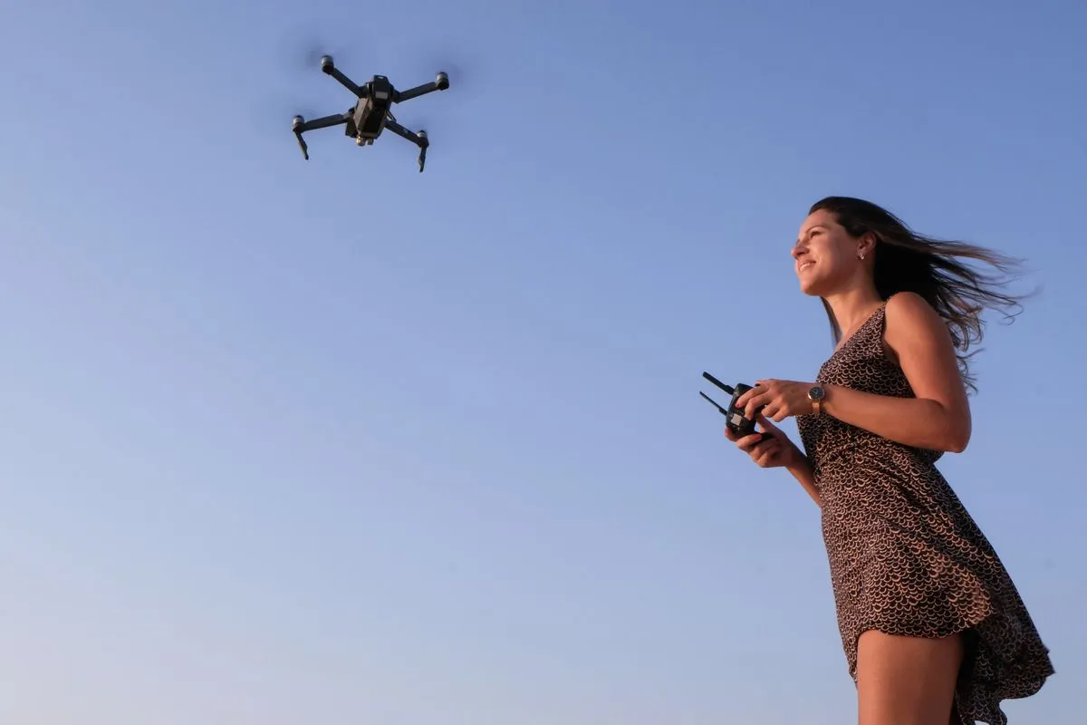 A woman in a patterned dress stands against a clear blue sky, holding a remote controller and watching the small drone she is flying above her