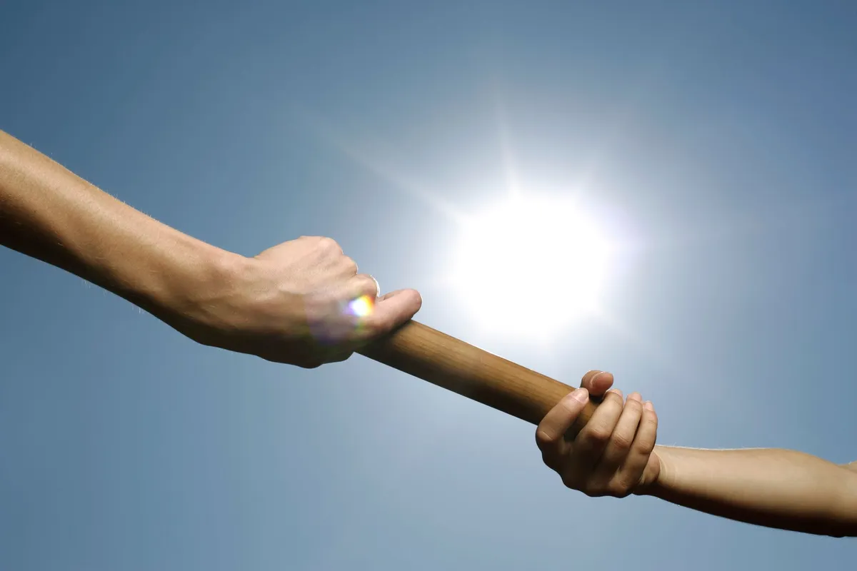 Two outstretched arms meet against a bright blue sky with the sun blazing behind them, one hand passing a wooden relay baton into the waiting grip of the other