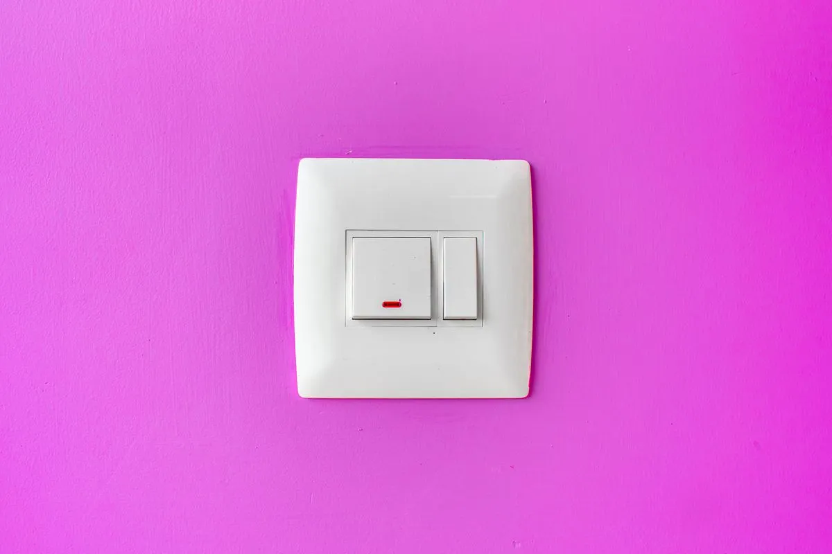 A white light switch with its small red indicator lit, mounted on a bright magenta wall