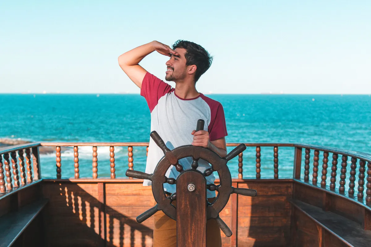 A man at a ship’s wheel shades his eyes and looks out over a bright turquoise sea, the wooden deck rail glowing in the sun