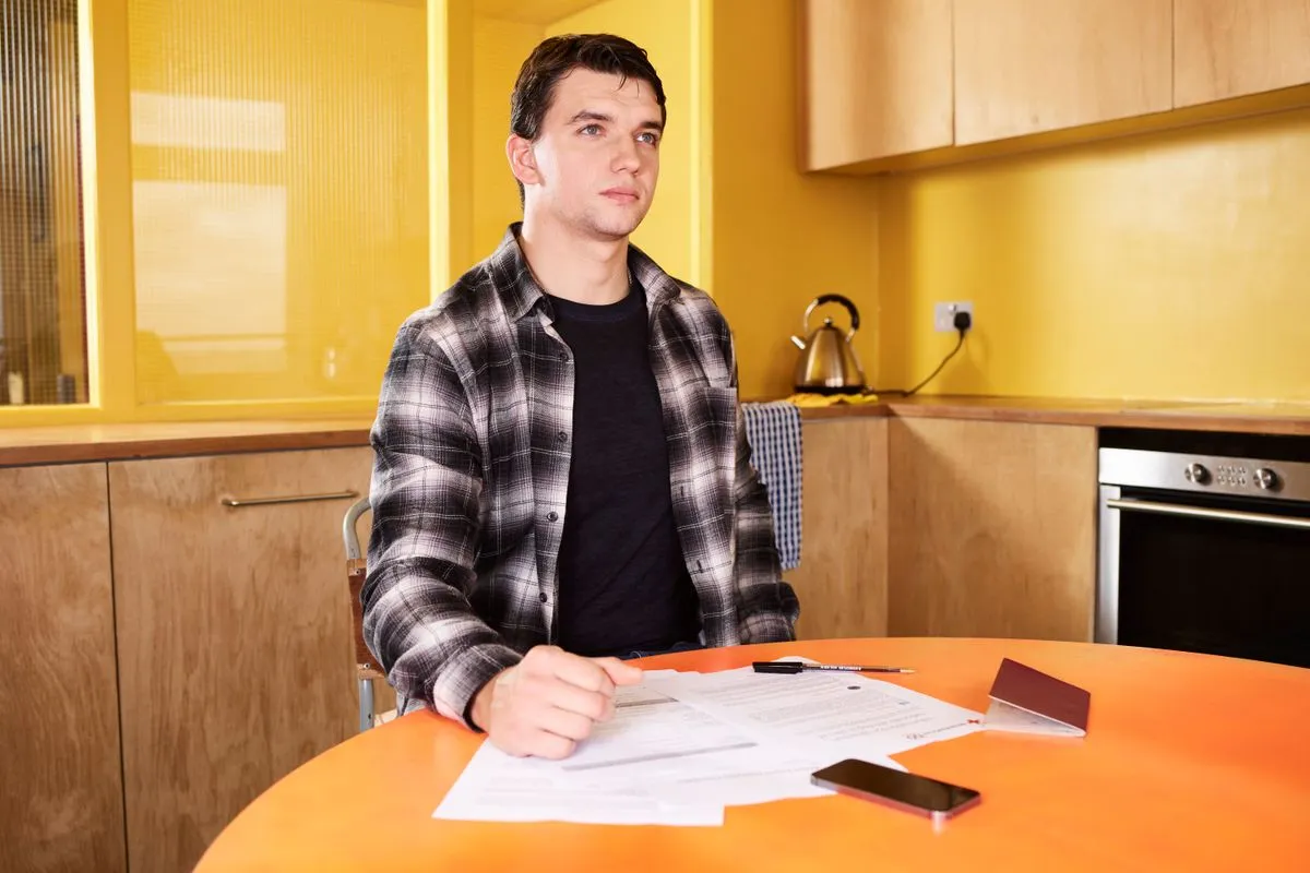 A man at a kitchen table reading a letter beside a laptop and a mobile phone