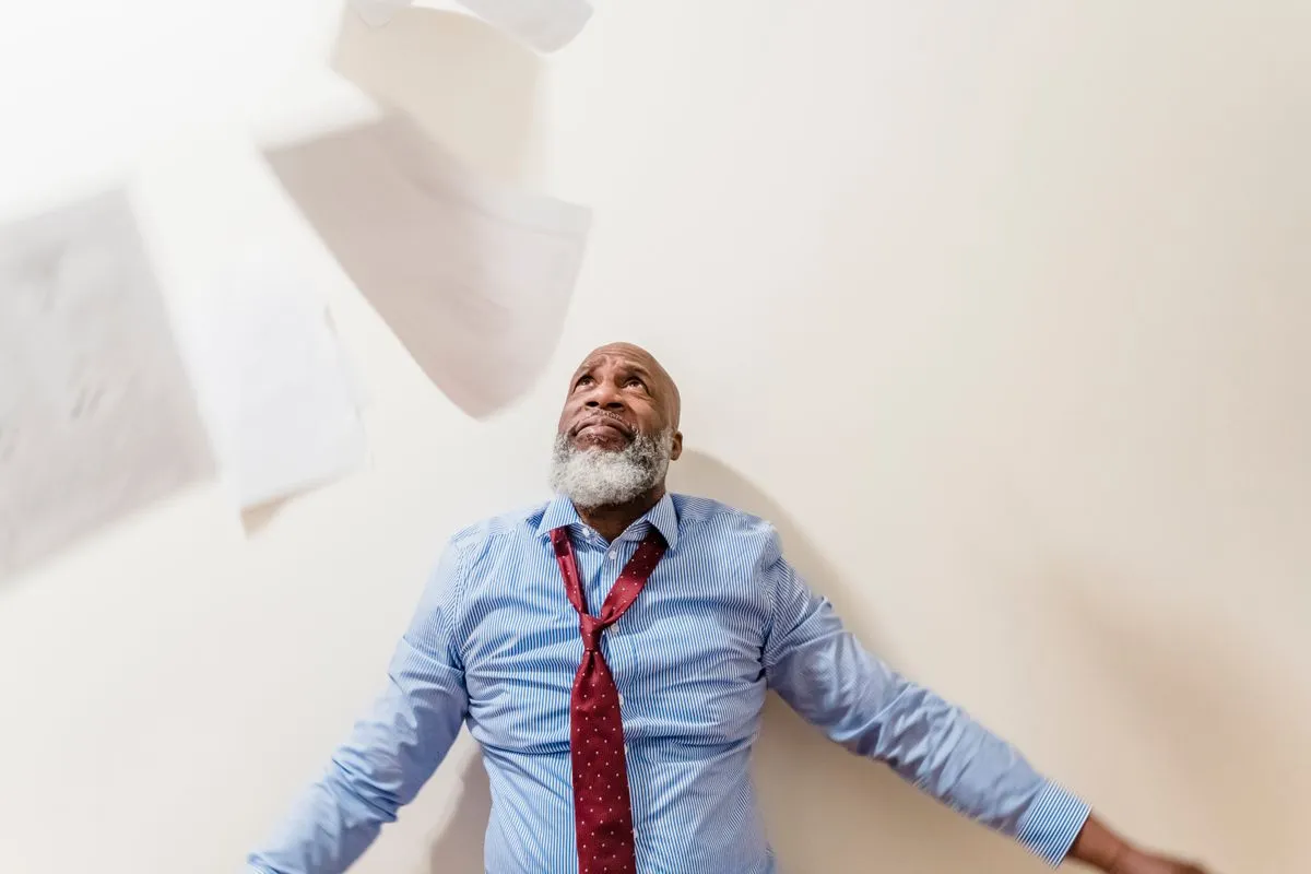 A grey bearded man in a blue striped shirt and loosened red tie stands against a cream wall, arms spread and eyebrows raised, looking up as sheets of paper tumble through the air around him