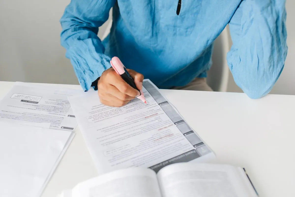 A person in a bright blue shirt sits at a white desk in daylight, running a pink highlighter along a few lines of a long printed document, with a second document and an open reference book beside it