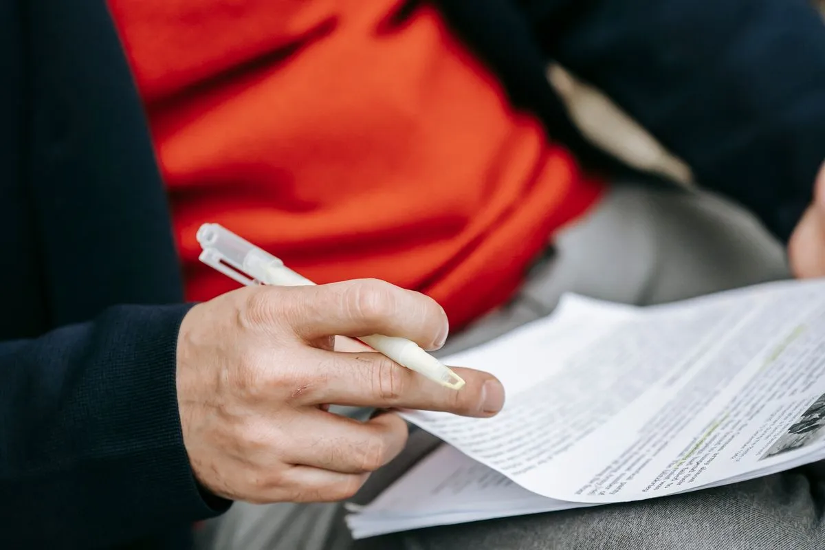 A person in a red jumper and navy jacket sits with a printed document on their knee, holding a highlighter pen over the lines they are checking
