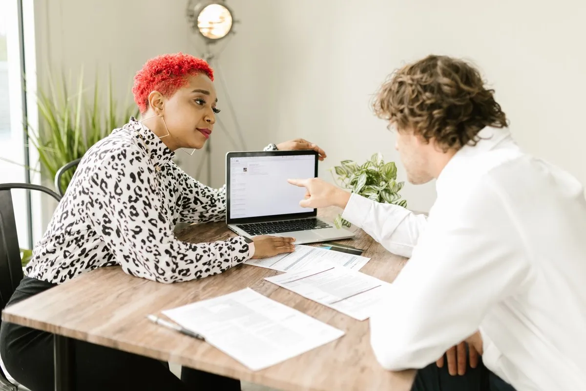 A woman with short bright red hair and a leopard print blouse sits at a wooden desk in a sunlit room while a colleague in a white shirt leans across and points at something on her laptop screen, printed pages spread on the desk between them